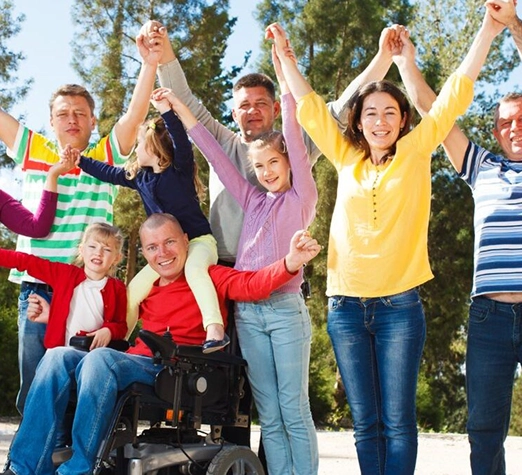 Services 3 - Reho Holistic Services Pty Ltd. A diverse group of adults and children, including a man in a wheelchair, stand outdoors holding hands and raising their arms, smiling joyfully with trees in the background.
