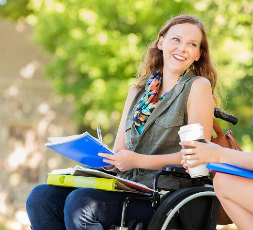 Services 2 - Reho Holistic Services Pty Ltd. A young woman in a wheelchair smiles while holding a coffee cup and notebook outdoors, talking with another person. The background is bright and green, suggesting a park or campus setting.