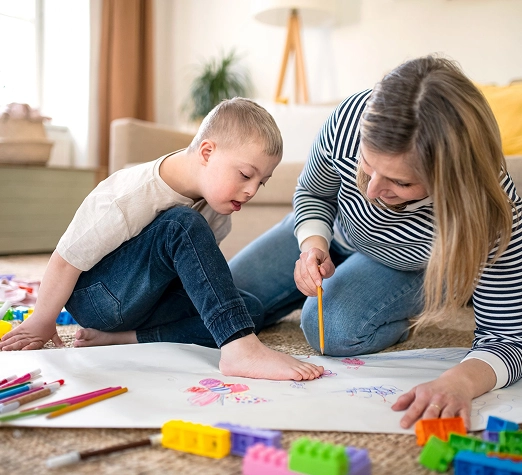 Services 6 - Reho Holistic Services Pty Ltd. A young boy and a woman sit on the floor, drawing together on a large sheet of paper with colored pencils. Colorful toy blocks are scattered nearby, and they appear focused and engaged in their artwork.