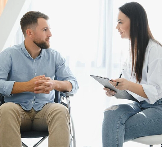 Services 9 - Reho Holistic Services Pty Ltd. A man in a wheelchair sits facing a woman holding a clipboard. They are engaged in conversation, both smiling, in a bright, casual indoor setting.