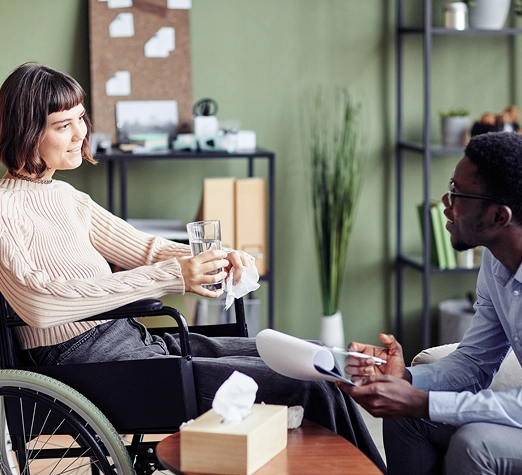 Services 1 - Reho Holistic Services Pty Ltd. A woman in a wheelchair holds a glass of water and tissue while talking to a man holding papers in an office setting, with shelves and a corkboard in the background.