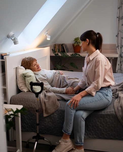 Home 1 - Reho Holistic Services Pty Ltd. A young woman sits on the edge of a bed, holding hands with an elderly woman who is lying down under a blanket in a cozy, well-lit room. A walking cane rests nearby.