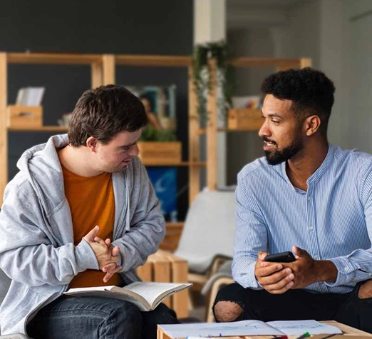Services 8 - Reho Holistic Services Pty Ltd. Two men are sitting indoors, engaged in conversation. One is holding an open book, and the other holds a smartphone. They appear to be having a friendly discussion in a cozy, well-lit room with shelves in the background.
