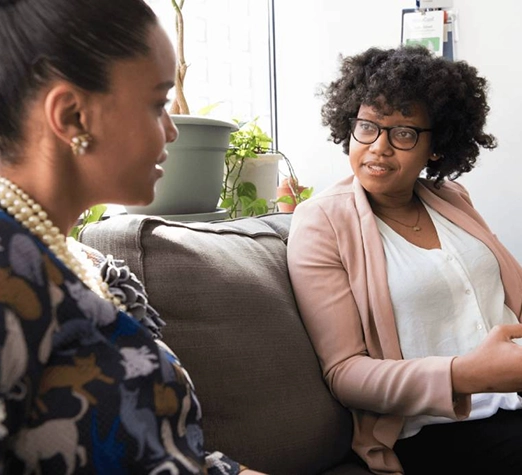 Services 7 - Reho Holistic Services Pty Ltd. Two women sit on a couch having a conversation. One wears glasses and a light pink blazer, while the other wears a patterned top and pearl necklace. There are plants and natural light in the background.