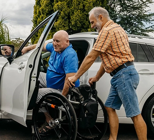 Services 5 - Reho Holistic Services Pty Ltd. An older man in a blue shirt transfers from a wheelchair into a white car with assistance from another older man in a plaid shirt and shorts, outdoors on a sunny day.